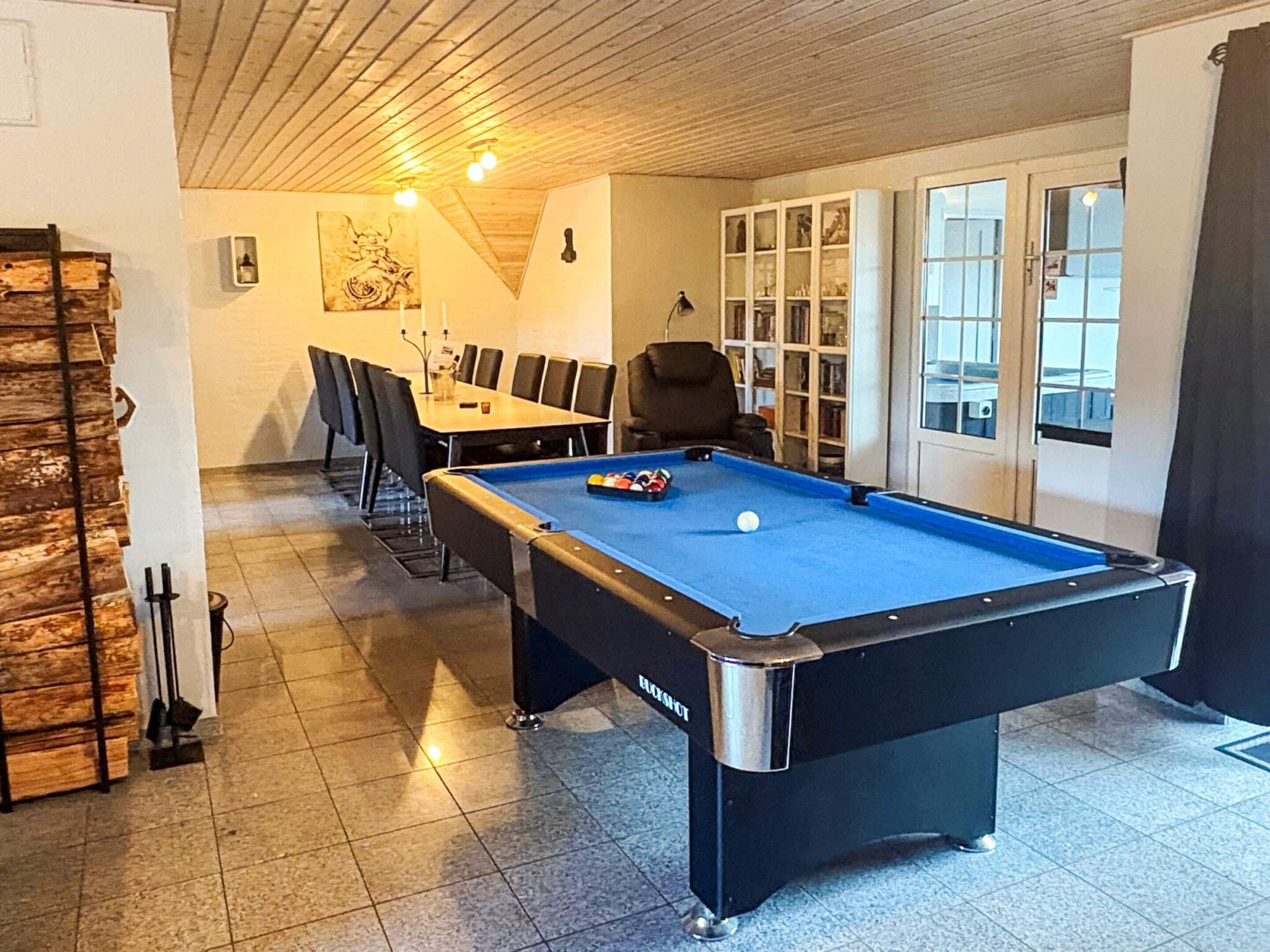 A pool table in a room with a wooden ceiling, a dining table with chairs, and a bookshelf.