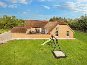 Une maison à toit de chaume avec une façade en briques, une terrasse avec des meubles d’extérieur et une balançoire installée dans une cour verdoyante.