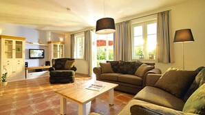 Un salon avec un canapé, un fauteuil et une table basse. On aperçoit un téléviseur et un meuble en arrière-plan.