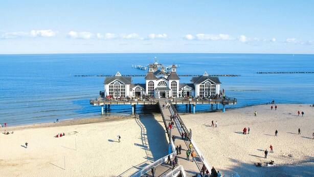 Une jetée mène à un bâtiment doté de nombreuses fenêtres et d’une tour centrale, qui surplombe une plage de sable où se trouvent des personnes.