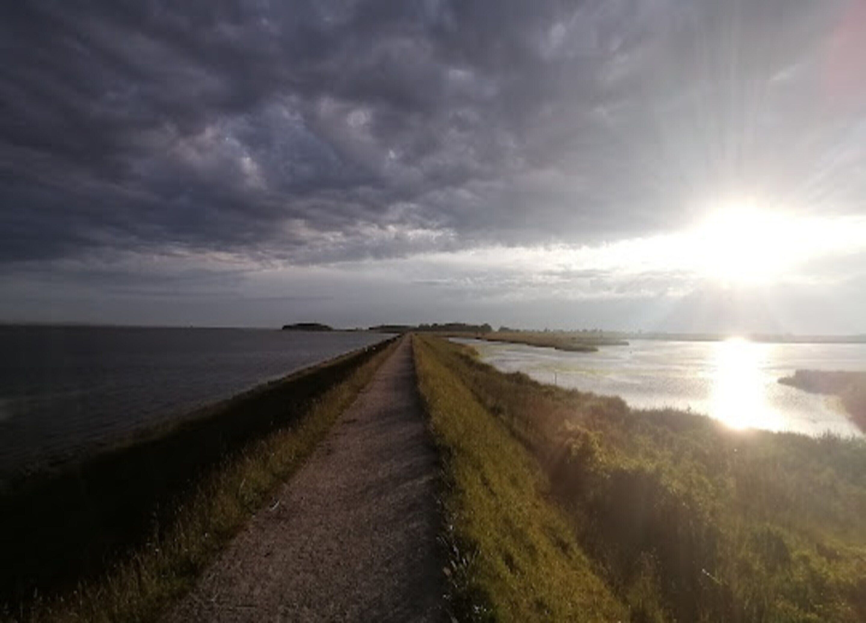 A coastal road with a clear path leading to the sea, under a cloudy sky with the sun visible on the horizon.
