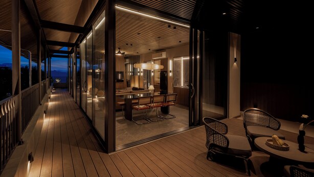 Un interior moderno con una terraza de madera, una zona de comedor con sillas y una mesa, y una cocina visible a través de puertas de vidrio.