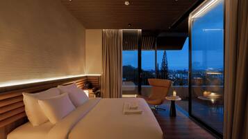 Una habitación de hotel moderna con una cama grande, un área de descanso con una silla y una mesa, y un balcón con vistas a la ciudad.