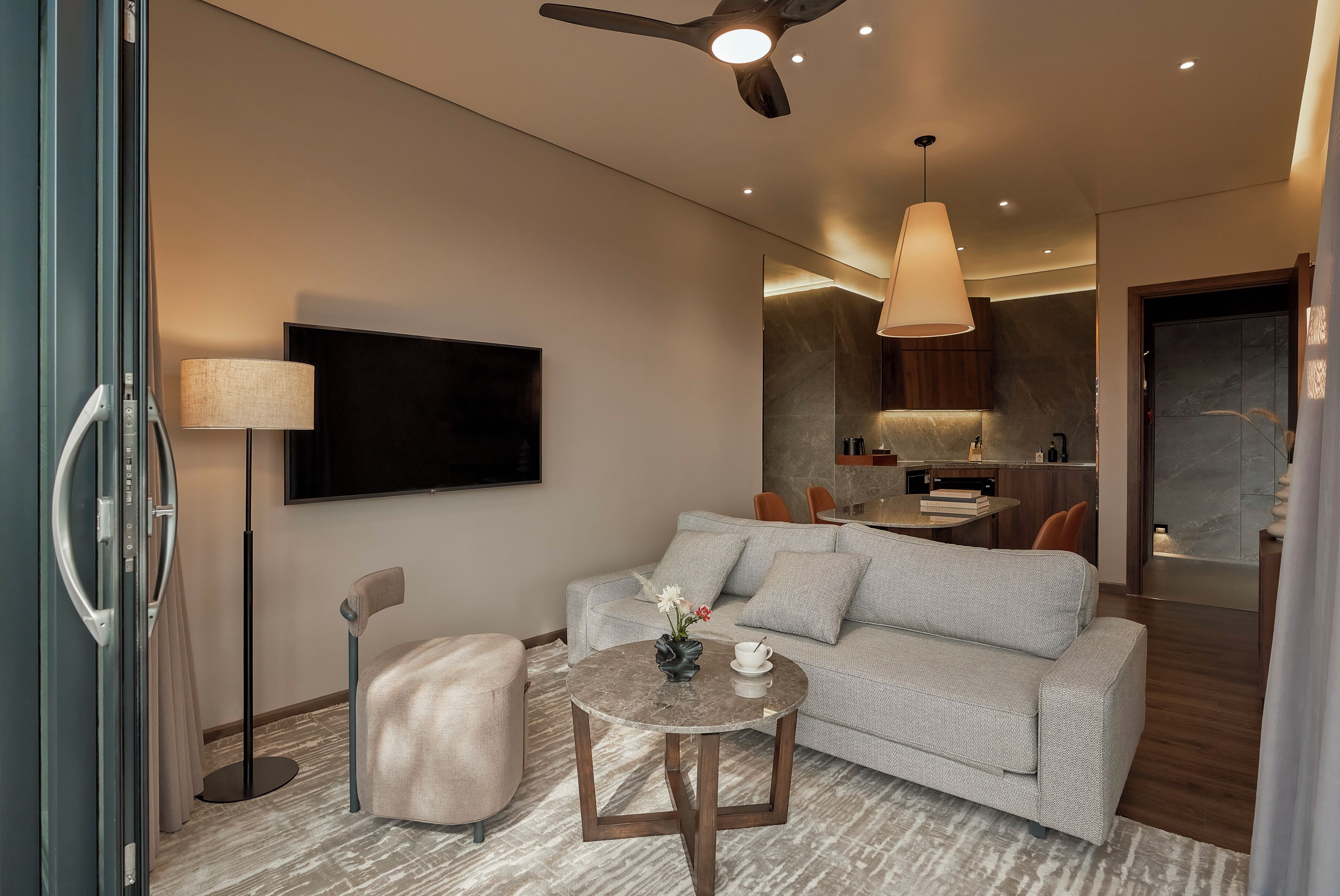 A modern living room with a grey sofa, a round coffee table, a floor lamp, and a flat-screen TV mounted on the wall.
