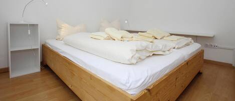 A wooden bed with white bedding in a room with white walls and a white shelf.