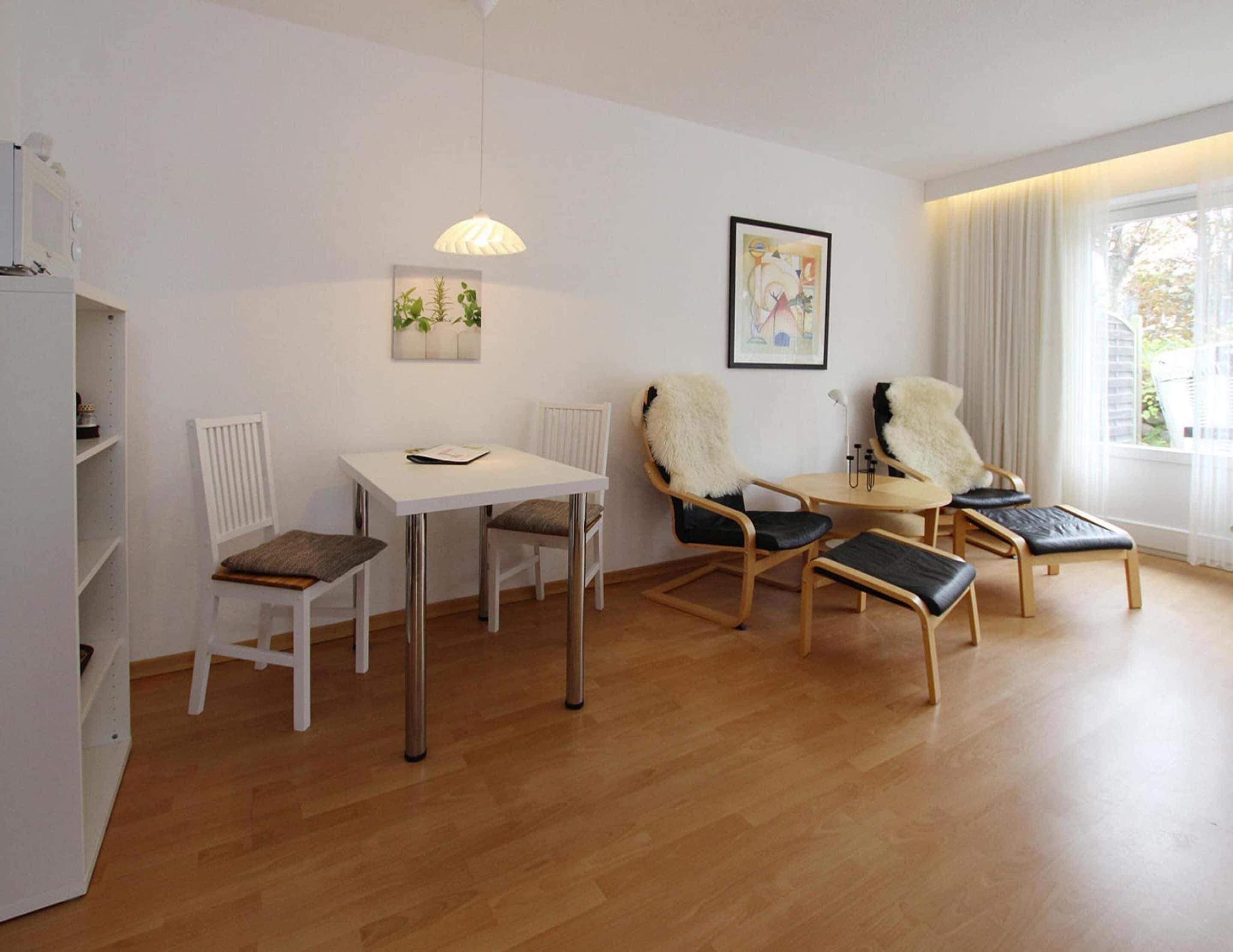Habitación moderna con una mesa blanca, dos sillas, una mesita redonda y un radiador. Hay una ventana con cortinas y un cuadro en la pared.