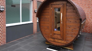 En tøndefarvet trækonstruktion med en dør, placeret på en belagt flade foran en murstensbygning med store vinduer.