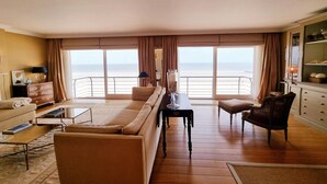 Ein geräumiges Wohnzimmer mit einer großen Couch, einem Couchtisch und einem Sessel. Ein großes Fenster bietet Blick auf den Ozean.