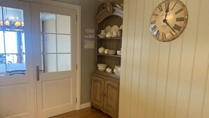 Ein Zimmer mit Holzboden, einer weißen Tür, einer an der Wand befestigten Uhr, einer hölzernen Bank und einem Regal mit Geschirr.