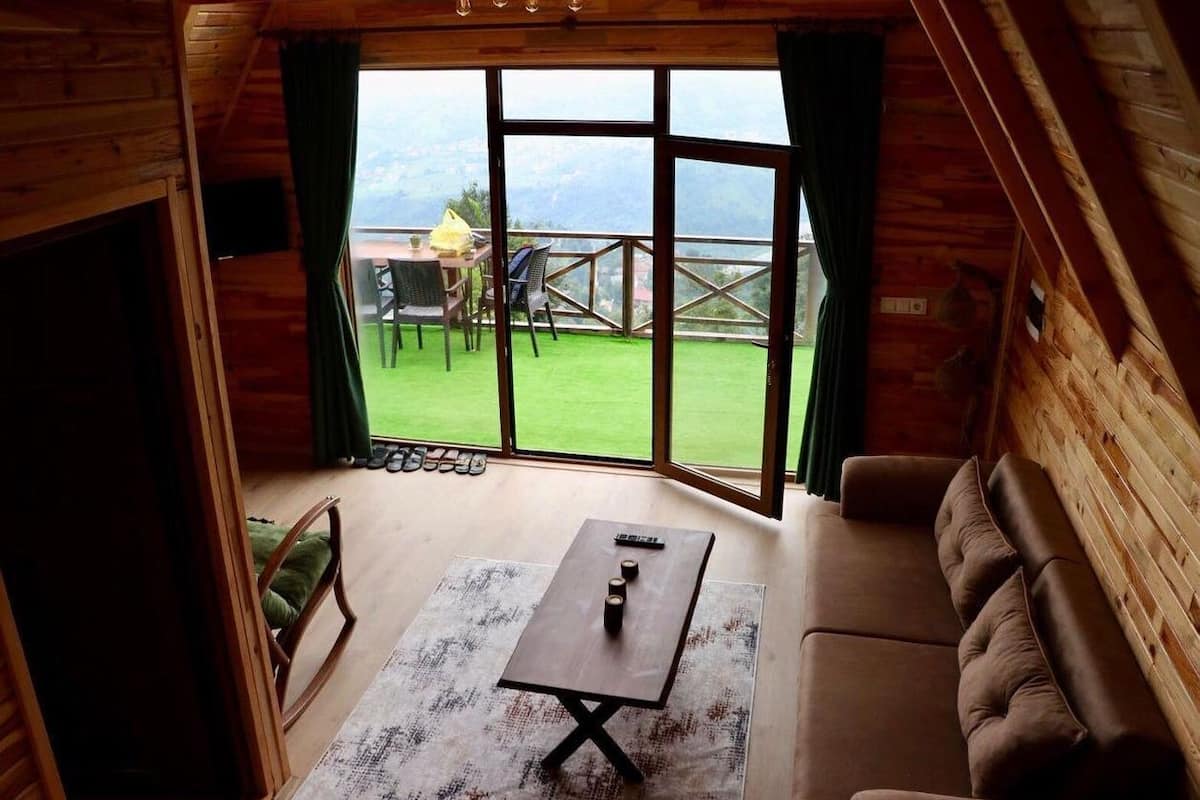 A cozy room with a wooden interior, a sofa, a table, and a view of a green lawn and mountains through large windows.