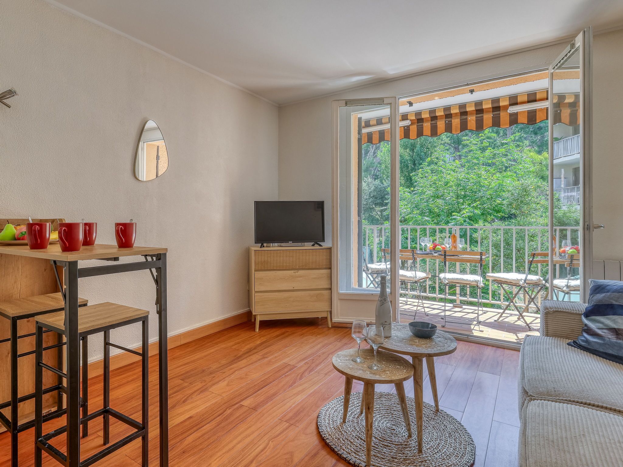 Una sala de estar moderna con piso de madera, un televisor sobre un mueble de madera, una zona de comedor con mesa y sillas, y un balcón con vistas a zonas verdes.