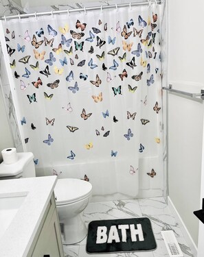 Kylpyhuone, jossa on perhoskuviollinen suihkuverho, wc-istuin, pesuallas ja musta kylpyhuoneen matto, jossa on teksti ”BATH”.