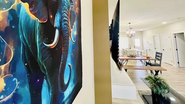 Ett modernt vardagsrum med en stor målning av en elefant, en platt-TV, en matplats med bord och stolar samt en kristallkrona.
