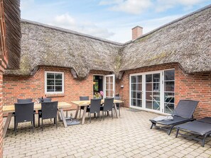 A brick building with a thatched roof patio area, featuring a table and chairs, a lounge chair, and a sliding glass door.