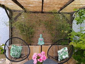 A covered outdoor seating area with two black wire chairs, each with a green and white patterned cushion. A small table with a vase of pink flowers is in the center.