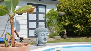 Un área de piscina tranquila al aire libre con una estatua de Buda, una planta en maceta y una casa de fondo.
