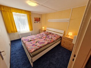 Una cama individual con edredón estampado, una mesita de noche con lámpara, un radiador, una ventana con cortinas amarillas y una estantería en la pared.