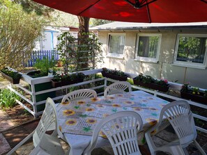Un patio al aire libre con una mesa preparada para cuatro personas, rodeado de sillas blancas, bajo un paraguas rojo.