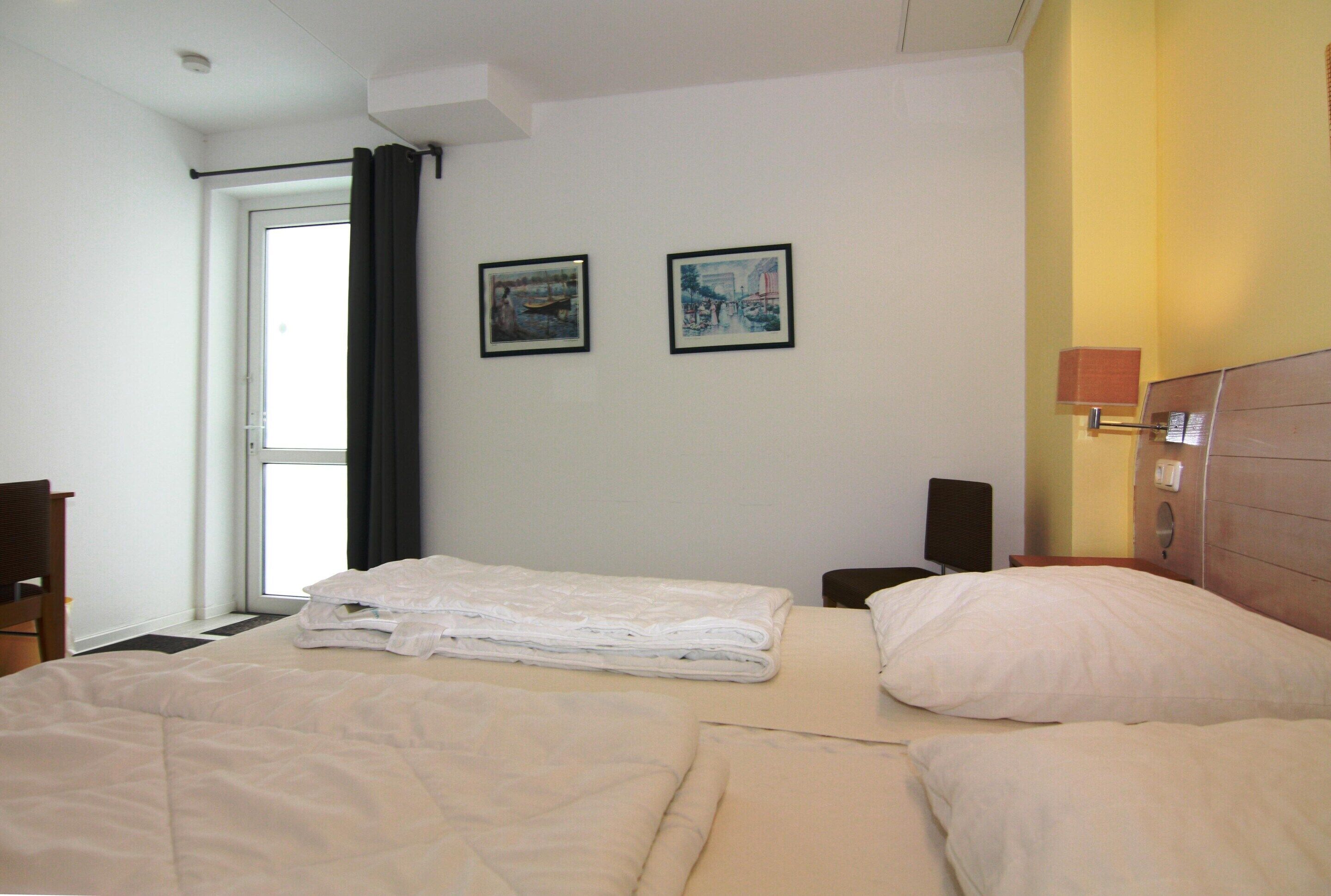 Ein Hotelzimmer mit zwei Betten, einem Fenster mit Vorhängen, zwei gerahmten Bildern an der Wand, einem Nachttisch mit Lampe und einem Holzkopfteil.