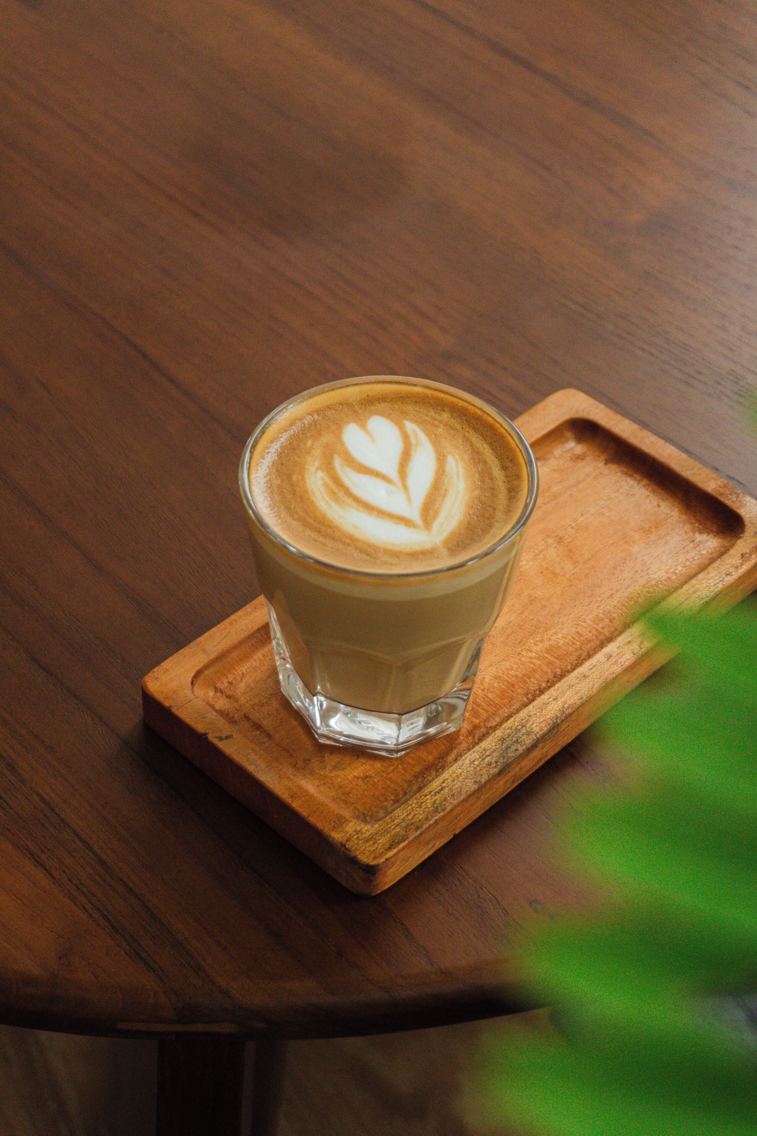 A glass of coffee with latte art on a wooden coaster on a table.