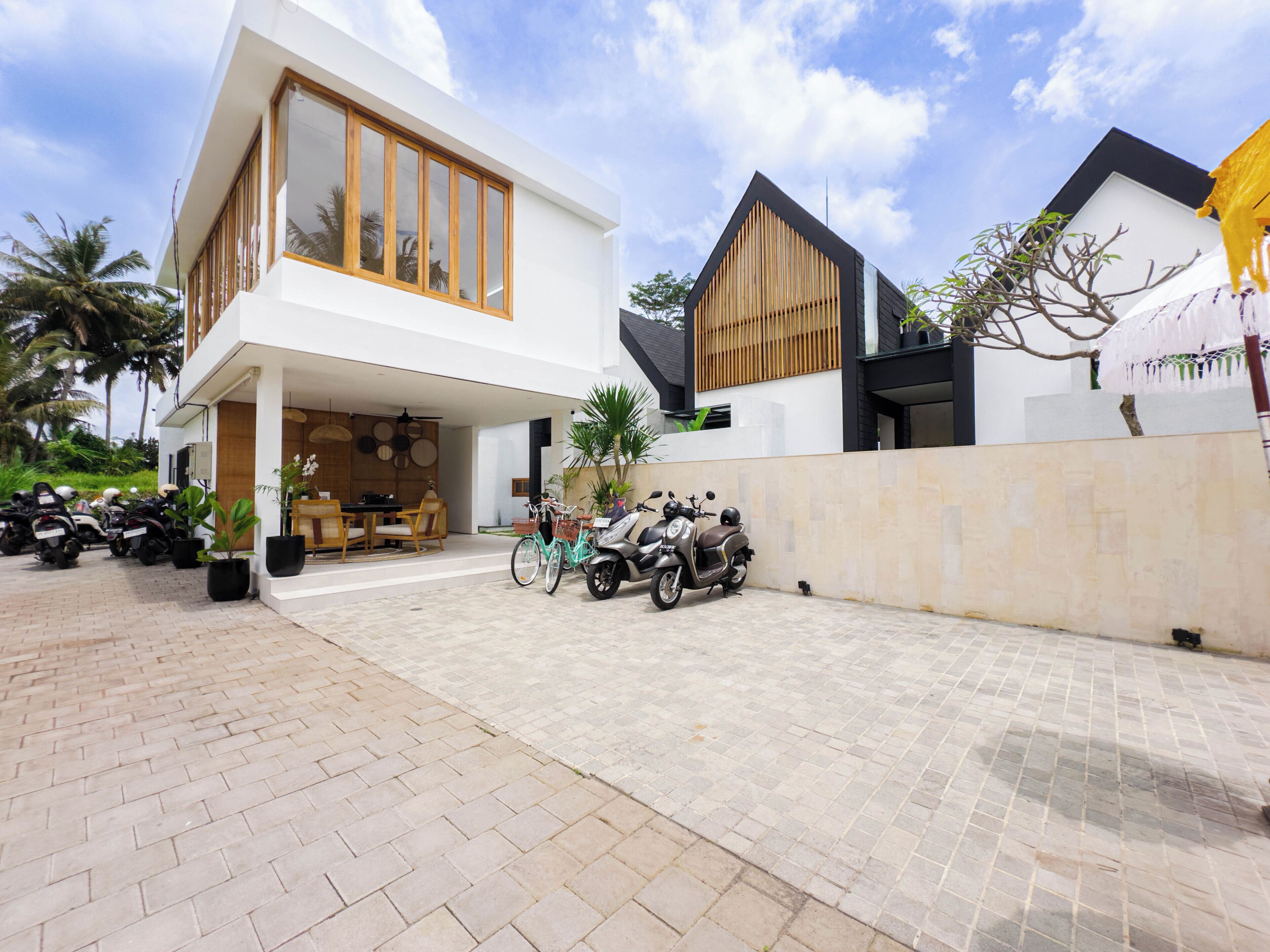 A modern two-story house with a covered patio, a dining area, and a parking area with several scooters and a bicycle.