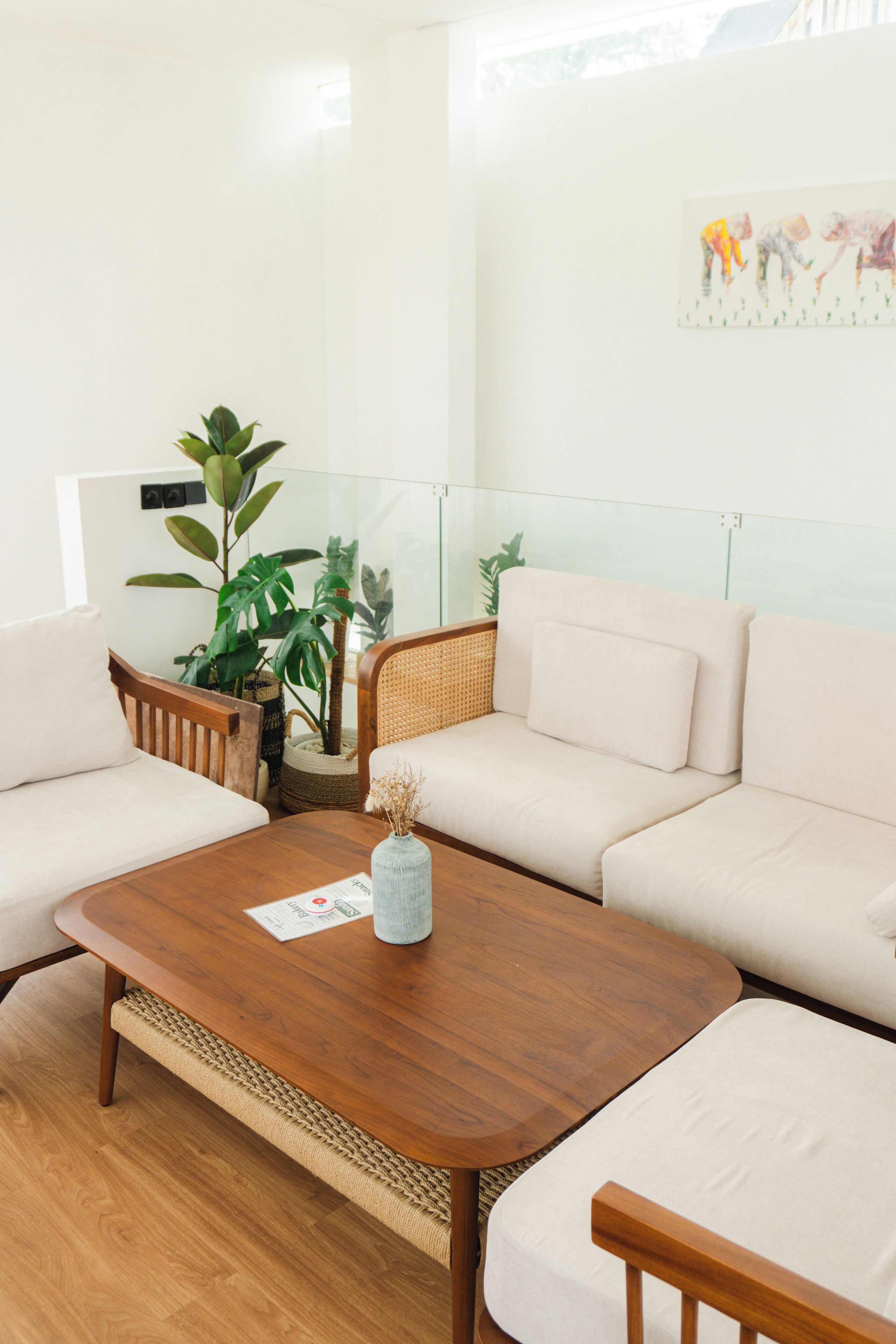 A modern living room with a wooden coffee table, a white sofa, and a rattan chair. There are potted plants and a painting on the wall.