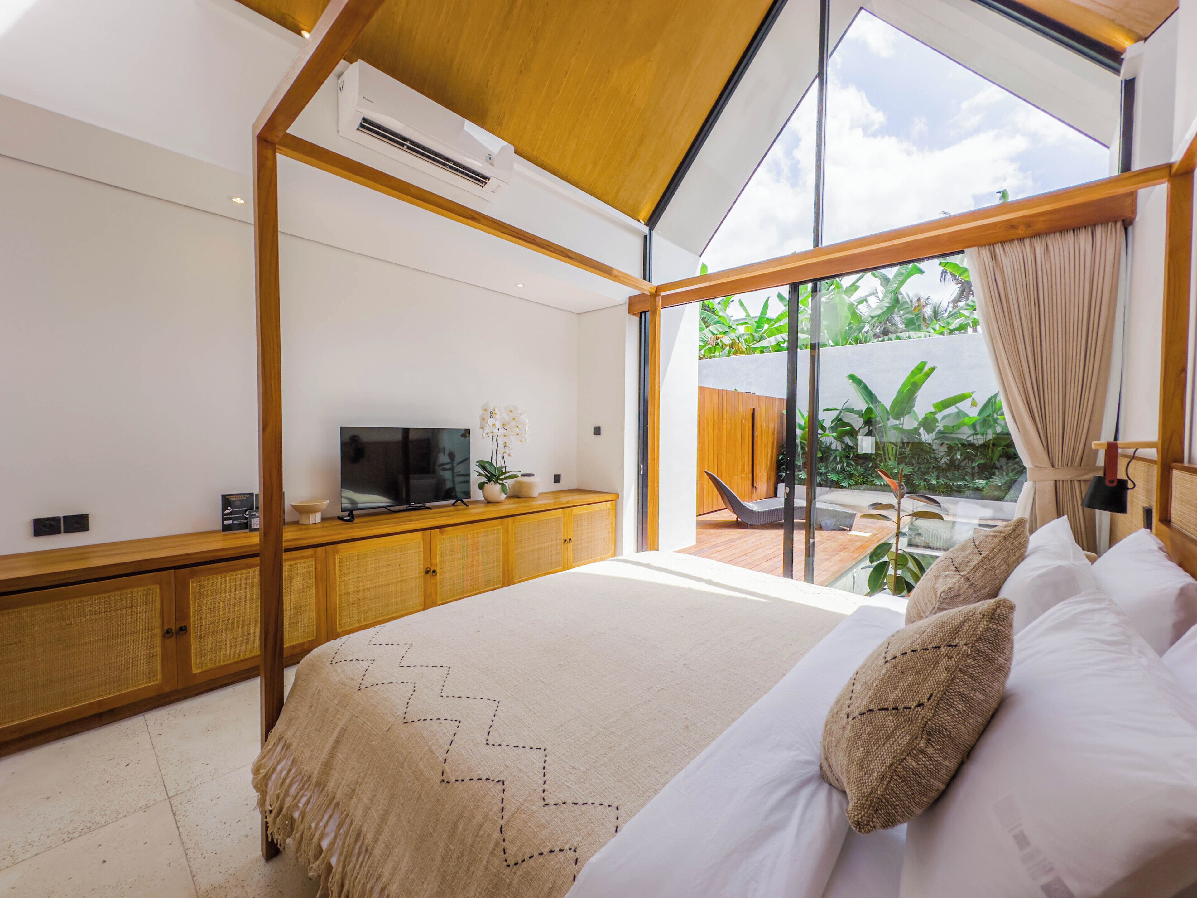 Un dormitorio moderno con una cama grande, un televisor sobre una consola de madera, y vistas a un jardín a través de puertas de cristal amplias.