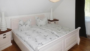 A bedroom with a white bed, floral bedding, a bedside table with a lamp, and a nightstand with a vase. - Ferienhof Landhaus Markus, Suderschwei (Stadland)