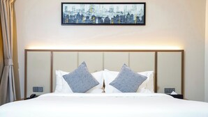Una cama bien hecha con sábanas blancas y dos almohadas grises, una obra de arte con un paisaje urbano enmarcado encima, y una mesita de noche con una lámpara y un teléfono.