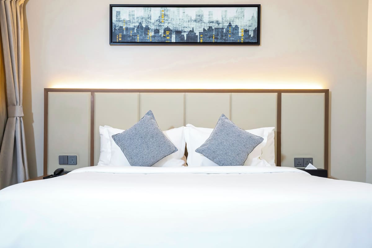 Een net opgemaakt bed met witte lakens en twee grijze kussens, een stadsgezicht in een lijst erboven, en een nachtkastje met een lamp en telefoon.