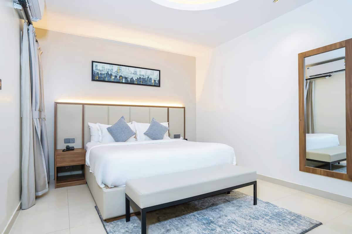 Een moderne hotelkamer met een groot bed, nachtkastjes, een bankje, stadsgezicht kunstwerk en een spiegel.