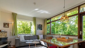 A modern living room with a sofa, a glass dining table, and a view of greenery through large windows.