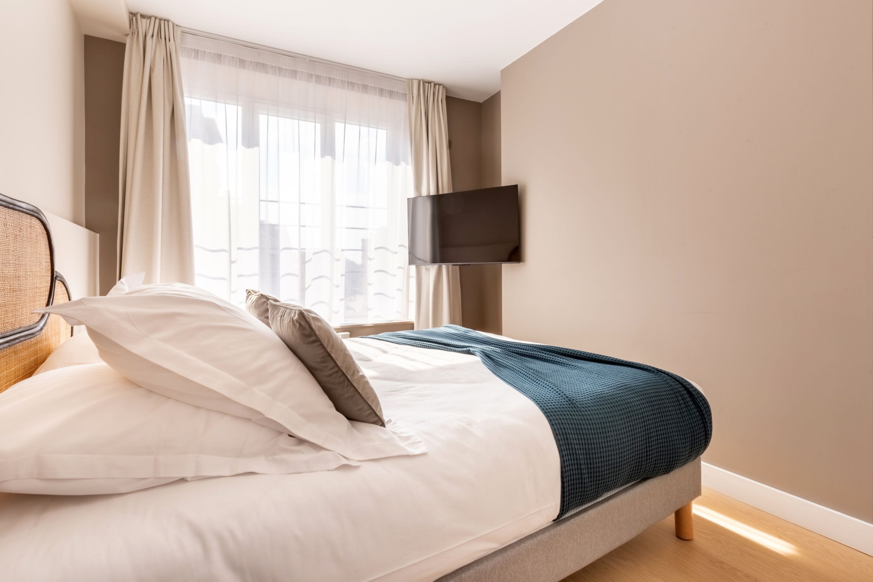 A neatly made bed with white and gray bedding, a teal throw blanket, and a flat-screen TV mounted on the wall.