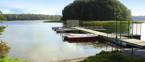 A calm lake with a dock extending into the water, surrounded by greenery and a forested hill.