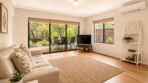 A living room with a sofa, TV, and a view of a garden through large windows. - The Aurora (Geographe)