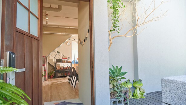 Une porte en bois donne accès à une pièce comprenant un coin repas, un escalier et un petit espace extérieur aménagé avec des plantes et des décorations à thème de grenouilles.