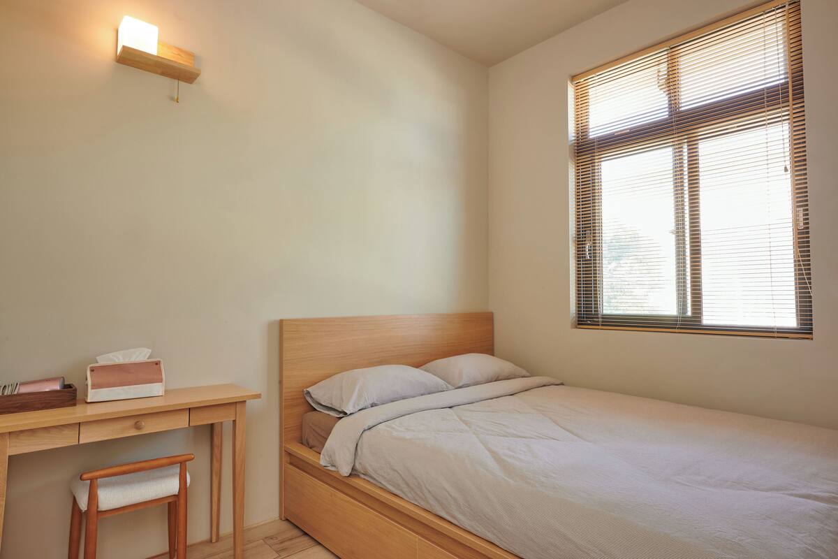 A bedroom with a single bed, a desk with a chair, a bedside table, and a window with blinds.