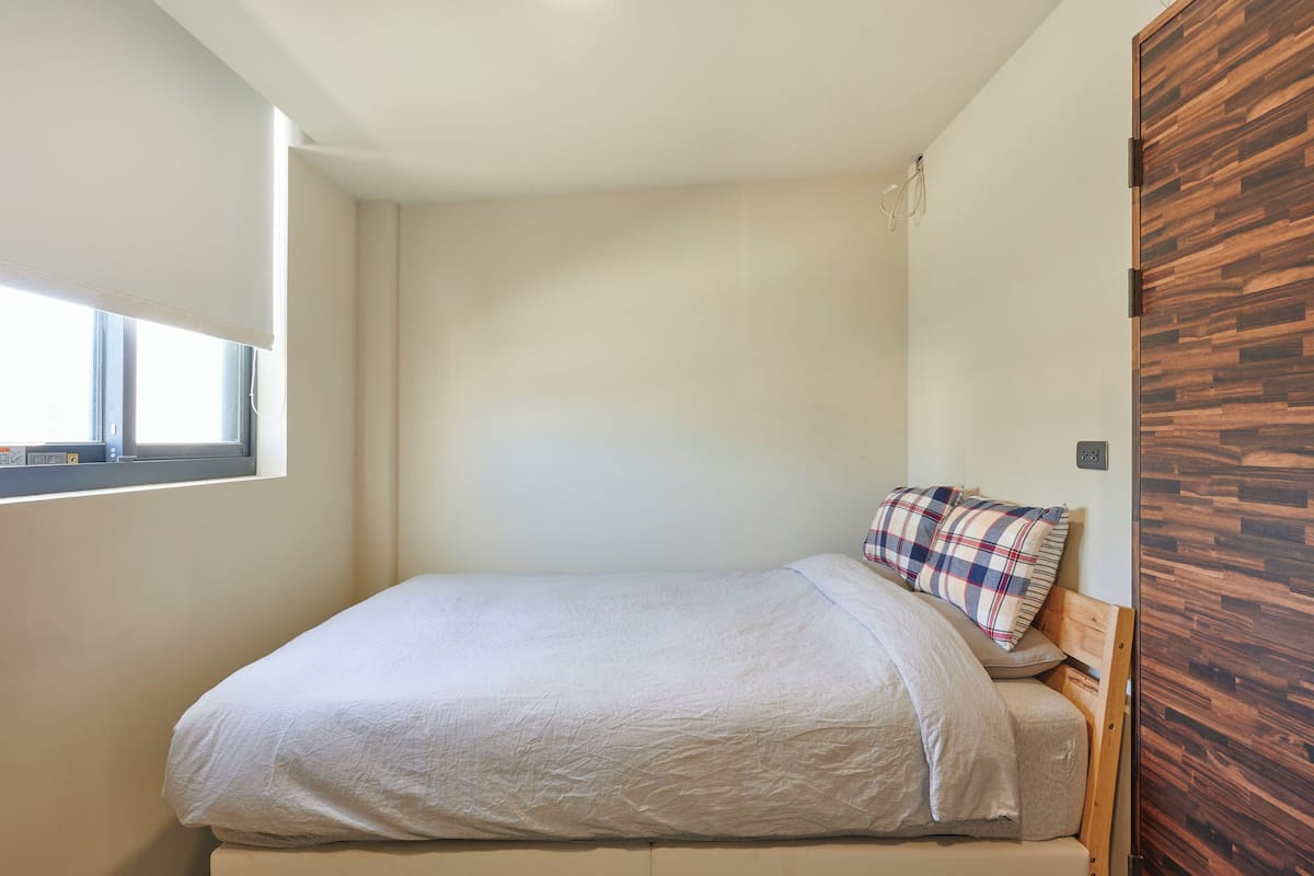 A small, neatly made bed with a plaid pillow and a plain white blanket in a room with a wooden accent wall and a window.
