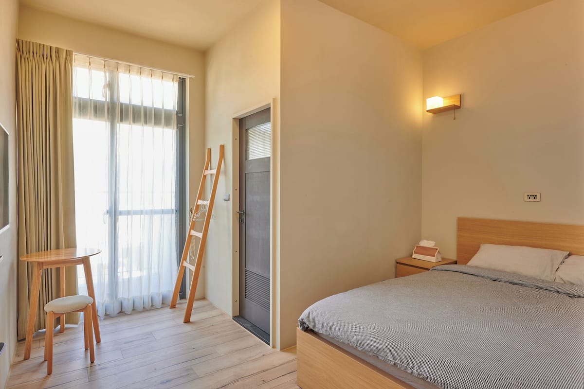 A hotel room with a bed, a bedside table, a chair, a wooden ladder, a window with curtains, and a wall-mounted light fixture.
