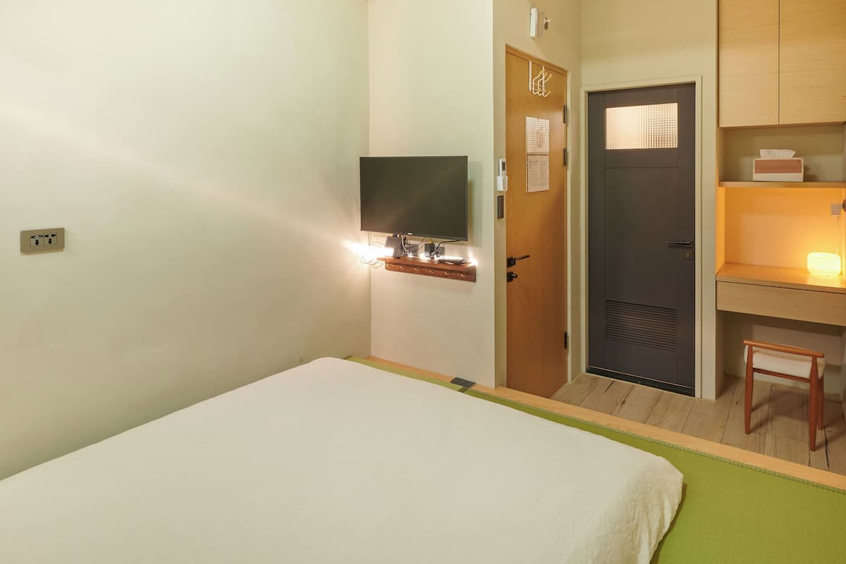 A hotel room with a bed, a TV mounted on the wall, a desk with a chair, and a door with a small window.
