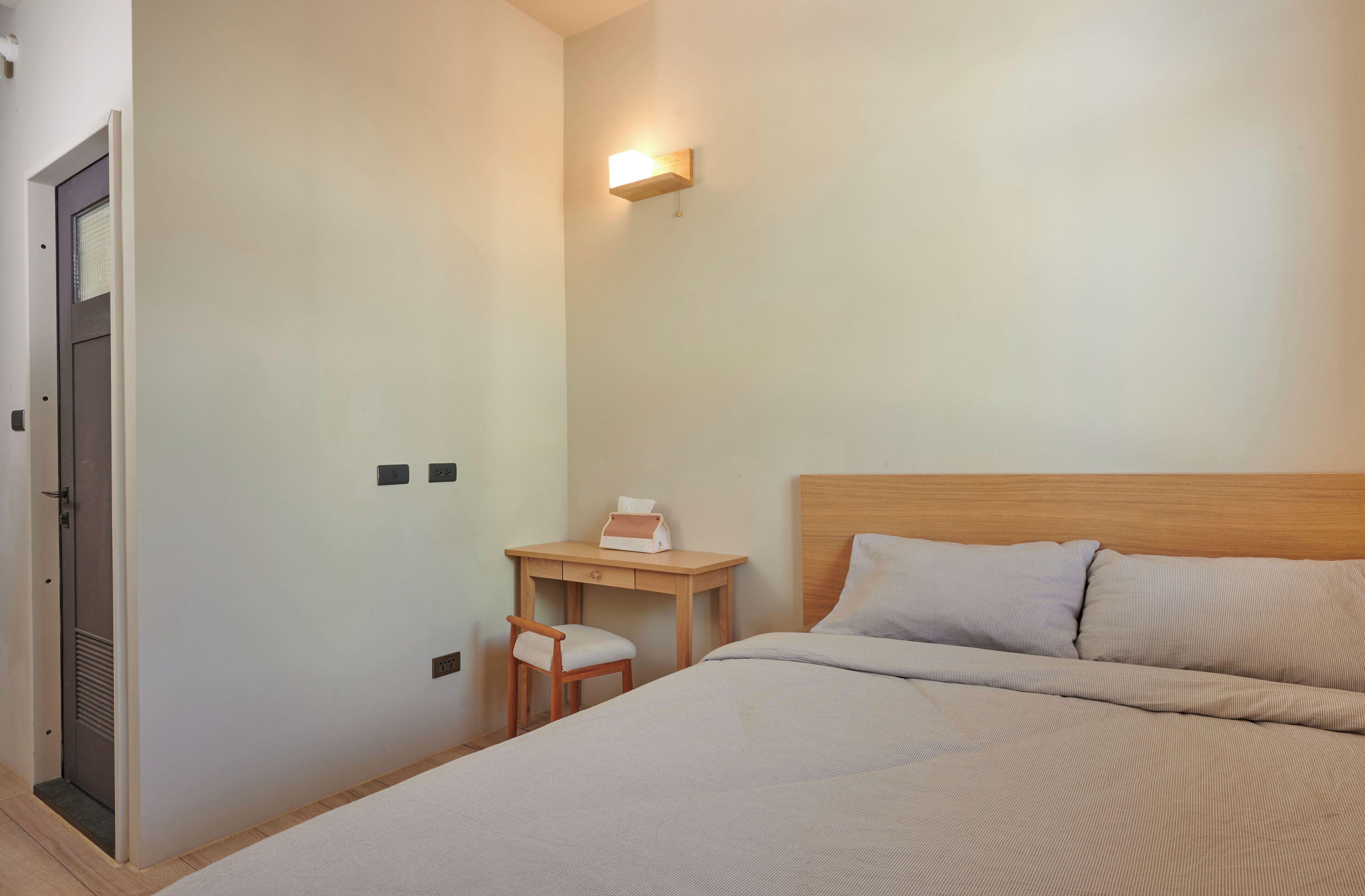 A hotel room with a bed, a desk with a chair, a nightstand, and a wall-mounted light.