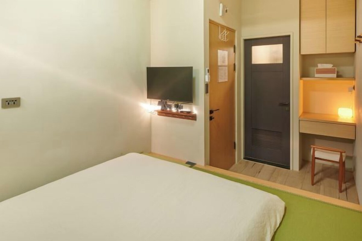 Ett hotellrum med en säng, ett skrivbord med en stol, en tv och en dörr med en spegel.