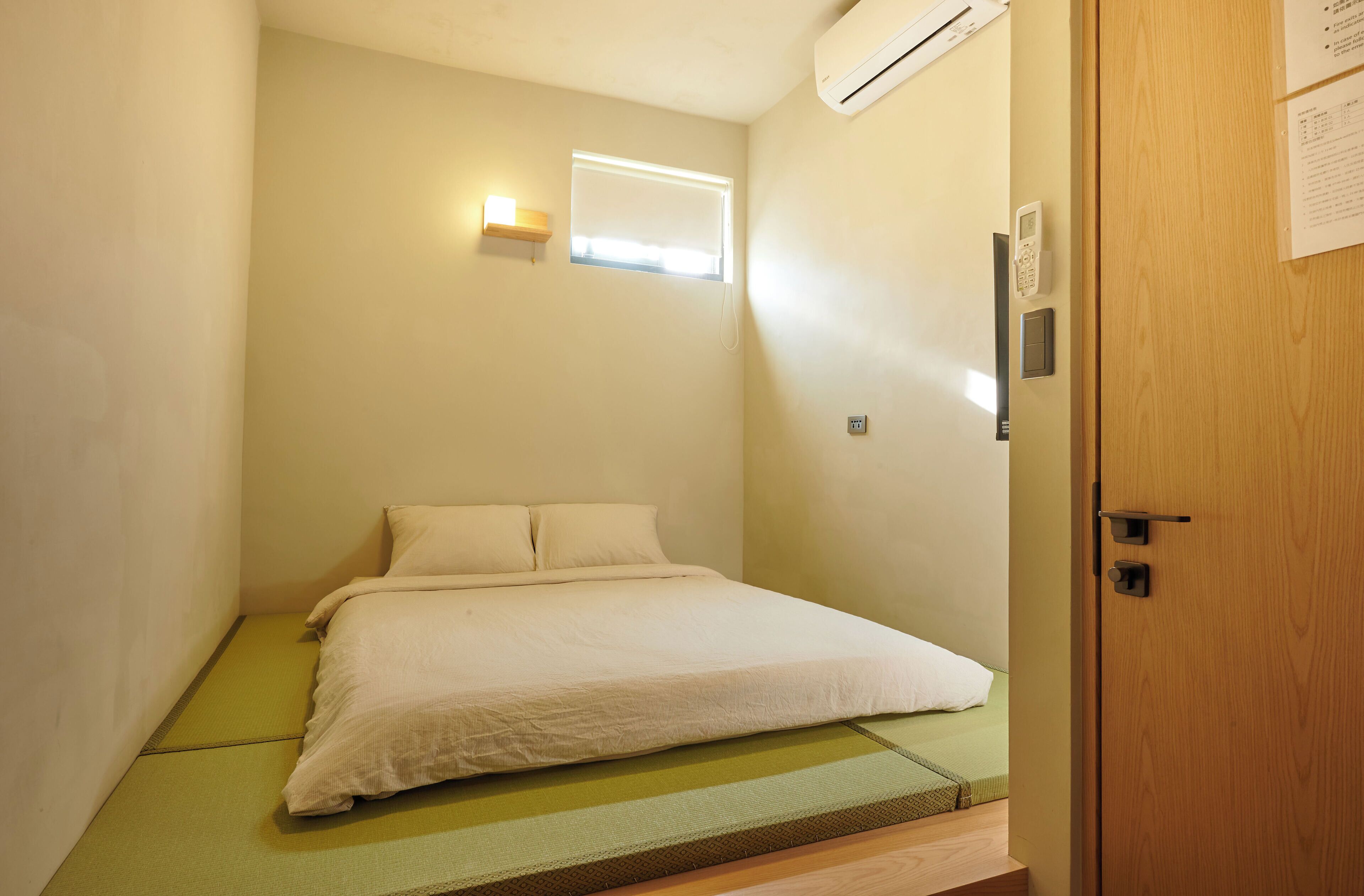 Une petite chambre à coucher, bien rangée, avec un lit, une fenêtre équipée de stores, un appareil de climatisation et une porte en bois.