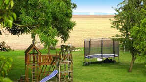 A playground with a slide, swings, and a trampoline in a grassy area with trees and a distant view. - Ferienhaus im Hafendorf Vieregge (Neuenkirchen)