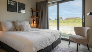 Een slaapkamer met een groot bed, een stoel, een raam met gordijnen en twee lijstjes aan de muur.