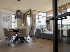 A modern dining area with a round table, chairs, and a sofa. Large windows provide a view of the outdoors. - Cottage in Wissenkerke Near Nature Reserve (Wissenkerke)