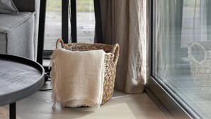 Een gezellige kamer met een grijze bank, een geweven mand en een raam met gordijnen.