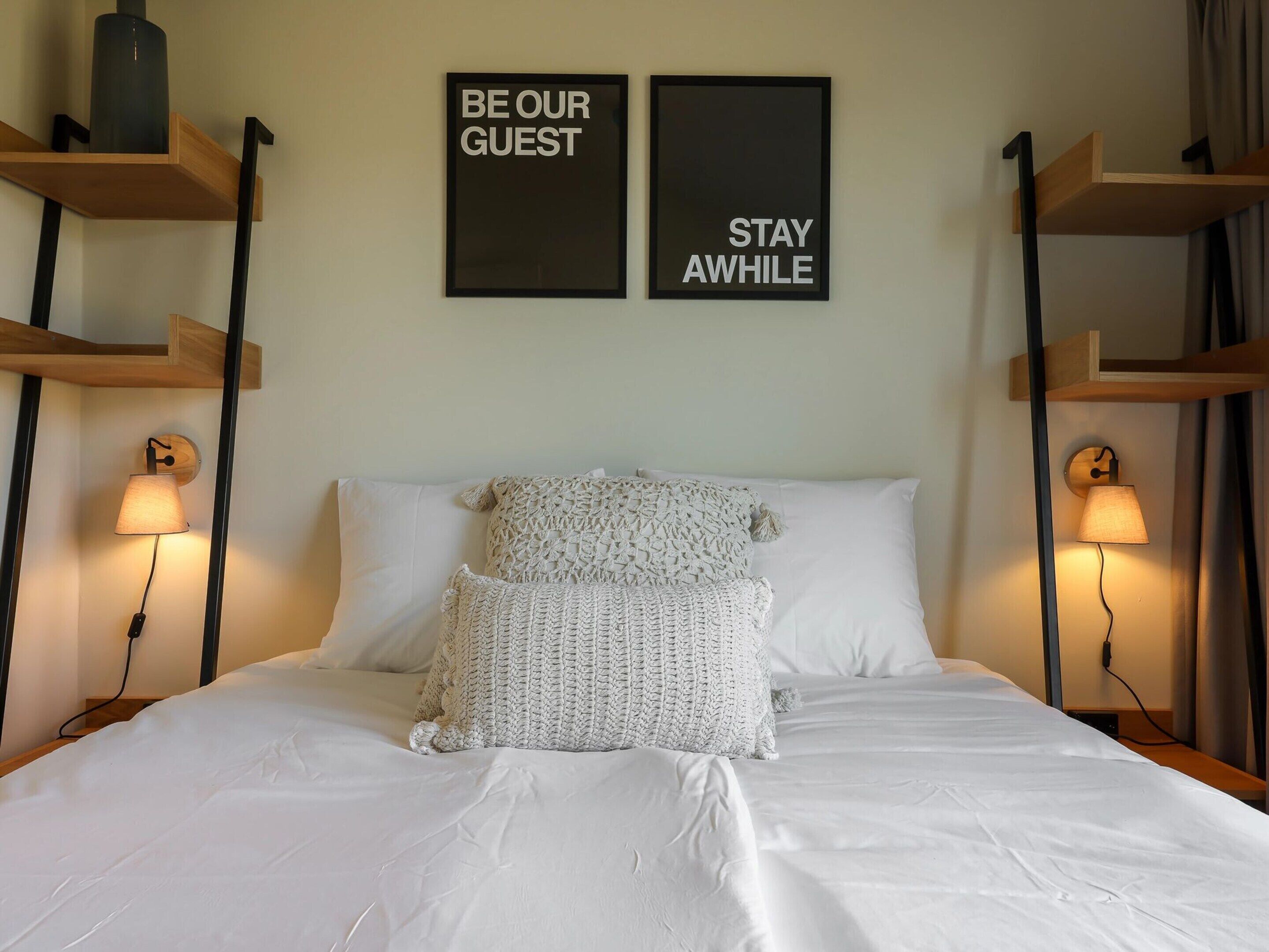 A hotel room with a bed, two wall-mounted signs reading "BE OUR GUEST" and "STAY AWhile", and two bedside lamps.