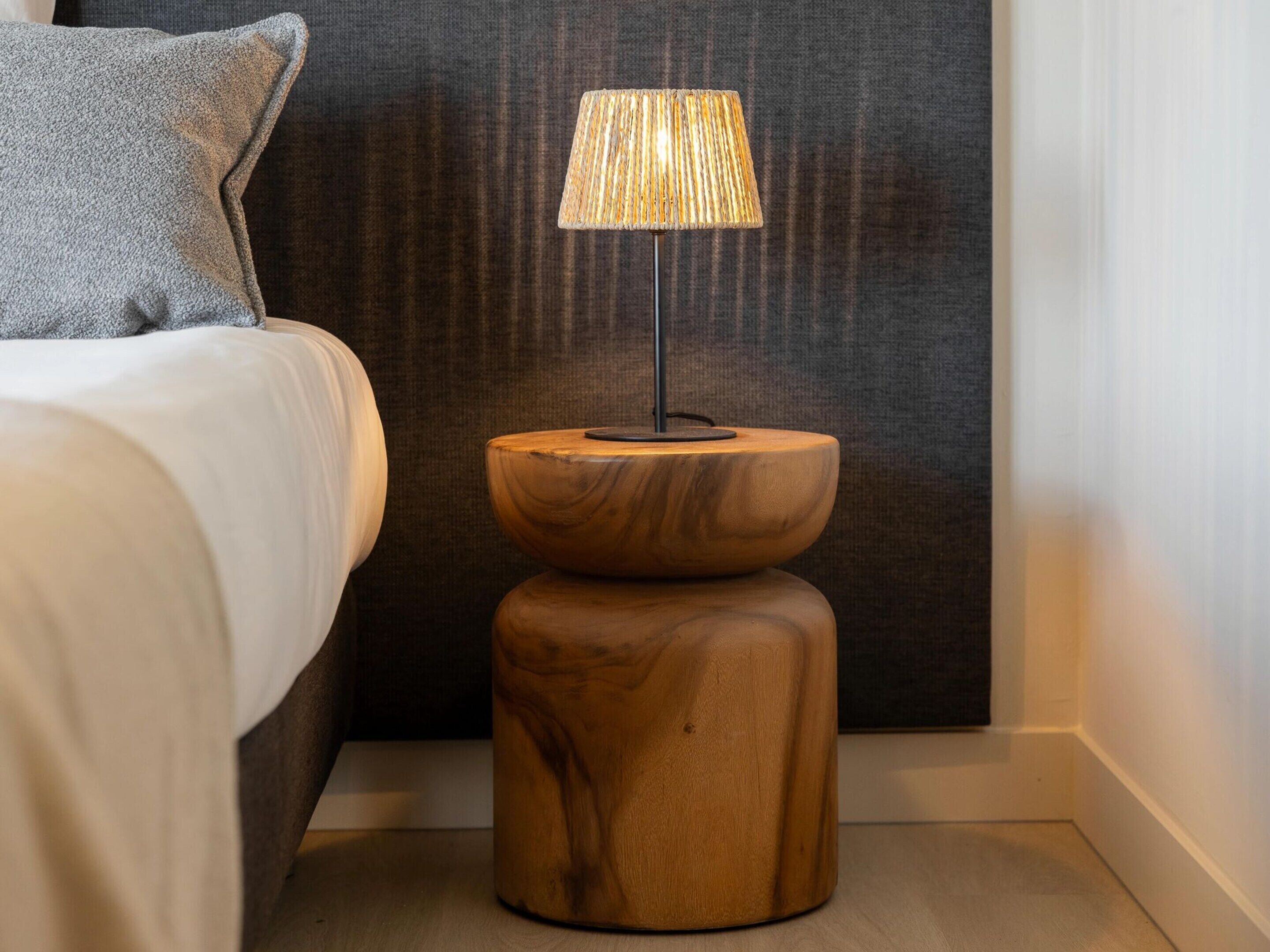 Een net opgemaakt bed met een grijs kussen, een houten nachtkastje met een lamp, en een donkere, gestructureerde wand.
