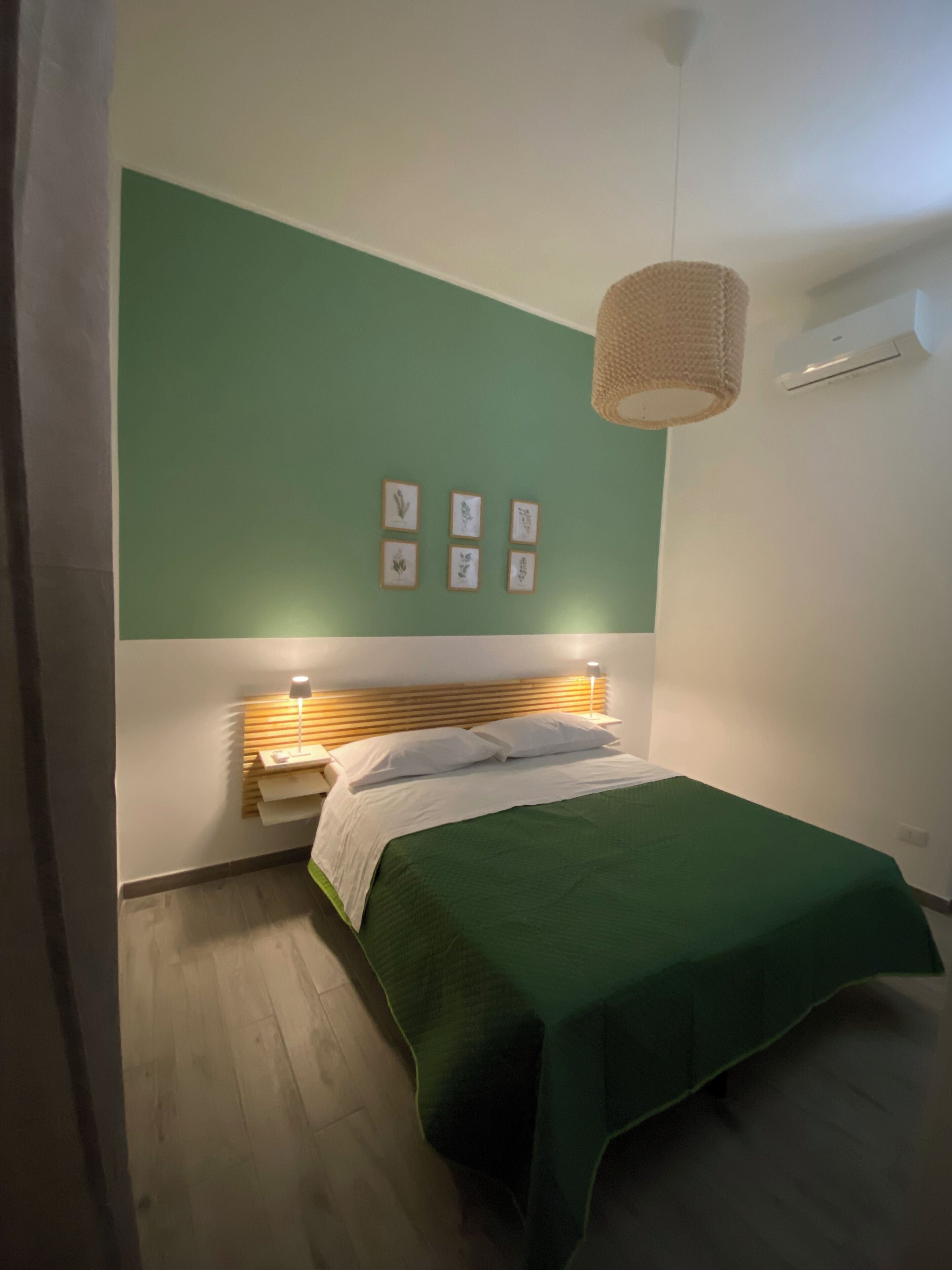 A bedroom with a green wall, a wooden headboard, a bed with a green bedspread, and framed botanical prints on the wall.